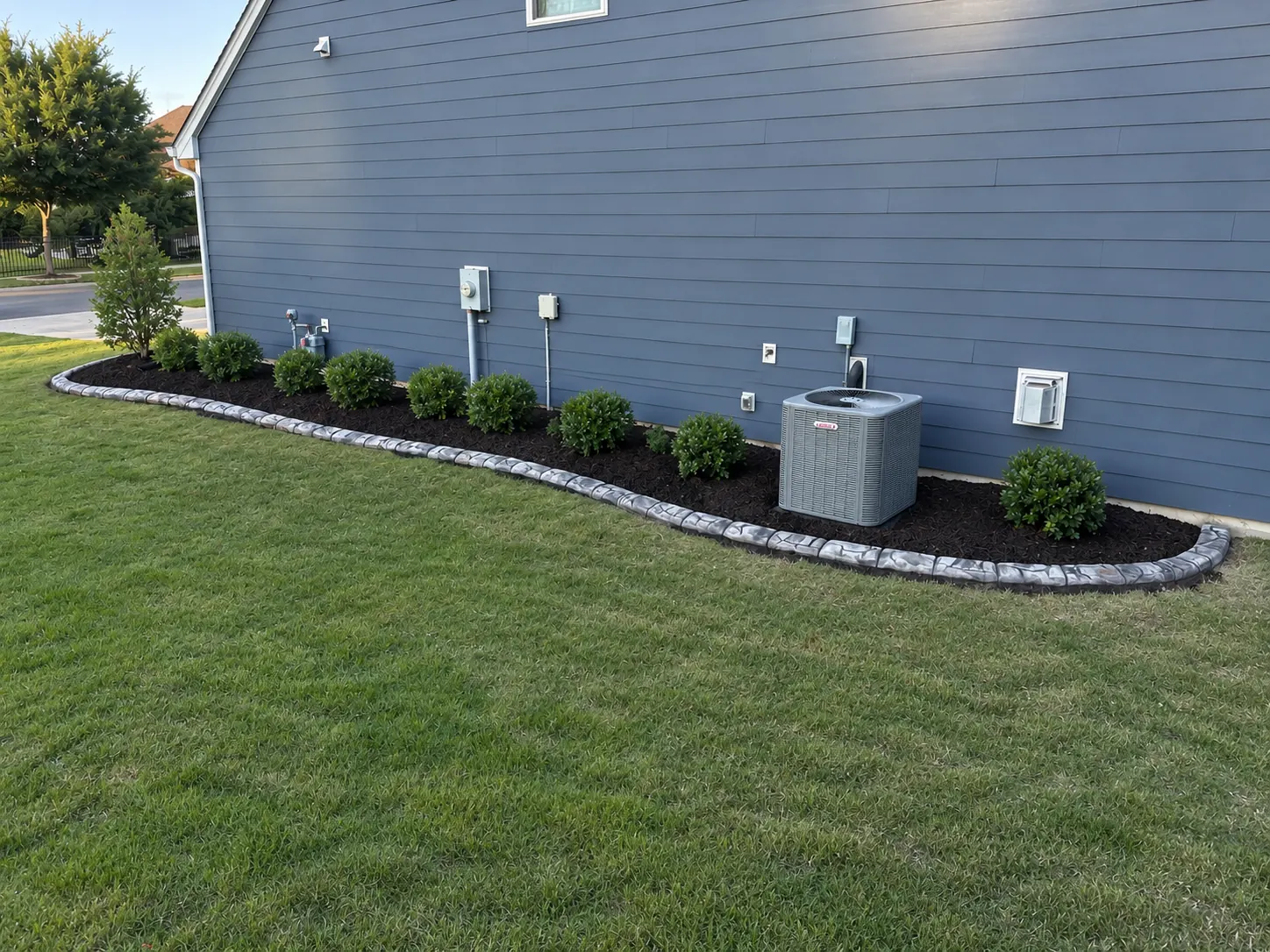 A long curved curb and mulched shrubs screening an air conditioner along a house wall